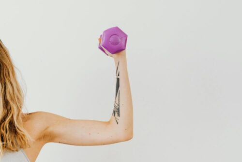 Close-up of a woman's tattooed arm lifting a purple dumbbell in a studio setting.